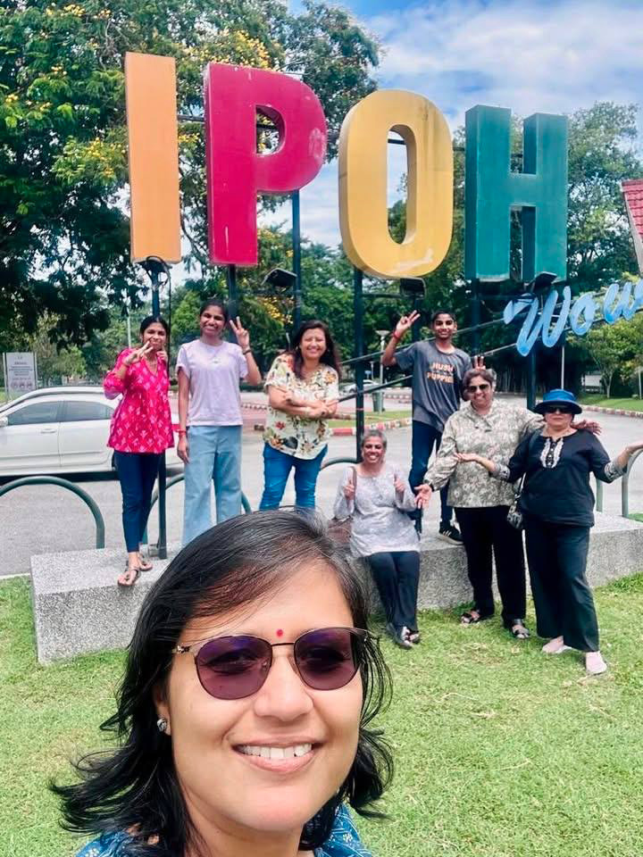A group of seven people smiling and posing with peace signs in front of large colorful letters spelling "IPOH" outdoors on a sunny day with trees and a car in the background.

Description generated by AI