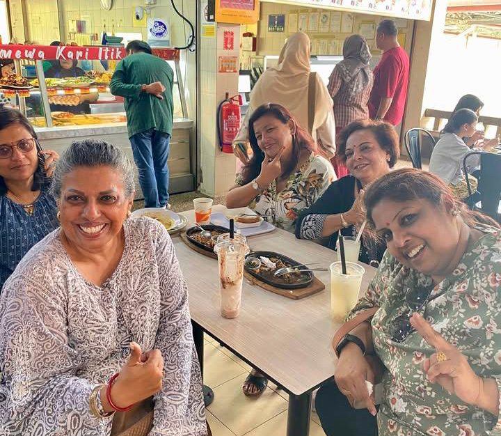 A group of five adults sitting around a table in a casual eatery smiling and posing for the camera with drinks and food trays on the table.
Description generated by AI