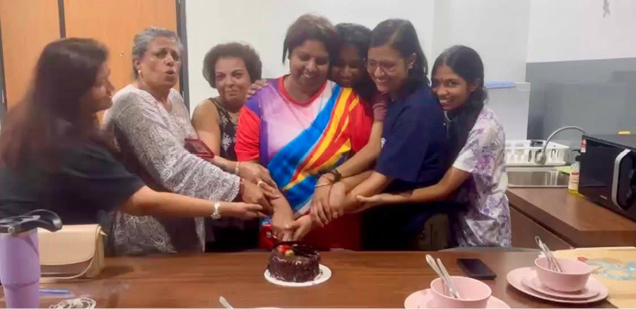 A group of seven people wearing colorful and casual clothing are gathered around a table smiling and cutting a chocolate cake together in a bright indoor setting.
Description generated by AI