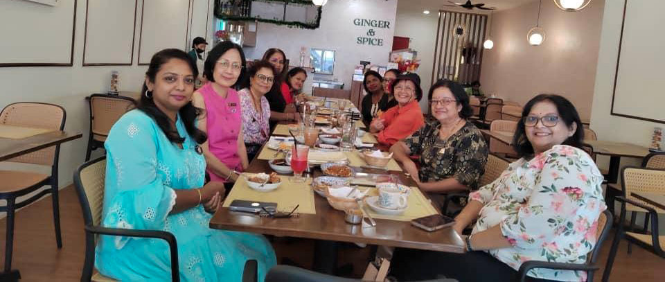 A group of people wearing colorful clothing are seated around a long table in a well-lit restaurant with modern decor, enjoying drinks and food.
Description generated by AI