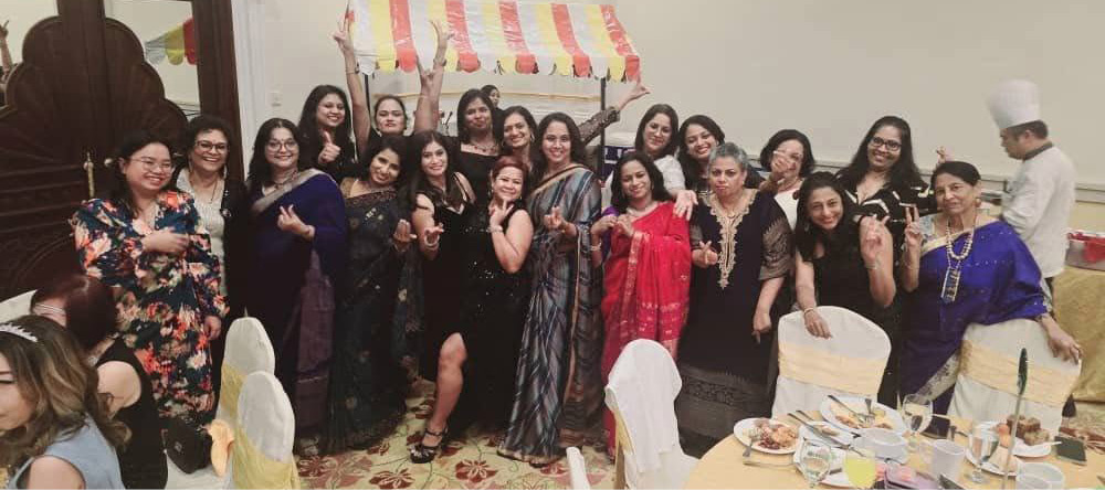 A group of adults dressed in formal and traditional attire are gathered indoors, smiling and posing for a celebratory group photo near decorated tables with food and drinks.

Description generated by AI
