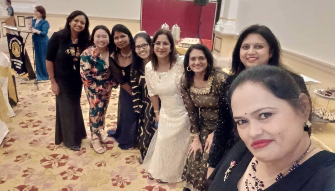 A group of seven adults dressed in colorful and elegant attire are smiling and posing closely together in a decorated indoor setting.

Description generated by AI