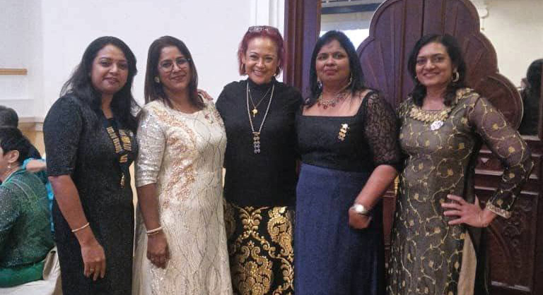 Five adults dressed in elegant traditional attire stand closely together indoors on a patterned carpet in front of a decorative wooden door.

Description generated by AI