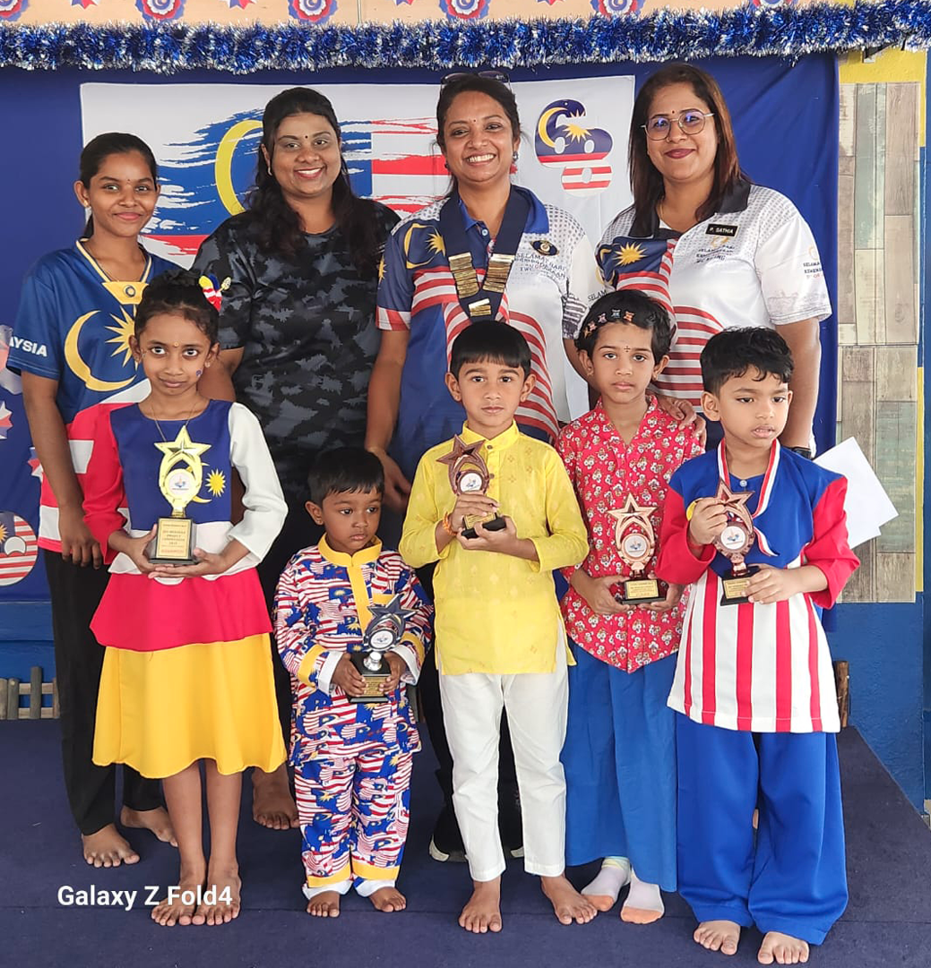 A group of adults and children dressed in colorful clothing stand together indoors with some children holding trophies and medals in front of a decorated backdrop.

Description generated by AI
