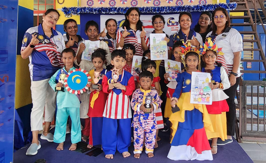 A group of children and adults dressed in colorful clothing are standing on a stage holding crafts and drawings, with a decorated backdrop and a staircase in the background.

Description generated by AI