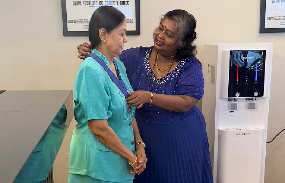 One person in a blue dress is placing a medal around the neck of another person wearing a teal outfit in an indoor setting with patterned floor tiles and framed posters on the wall.
Description generated by AI