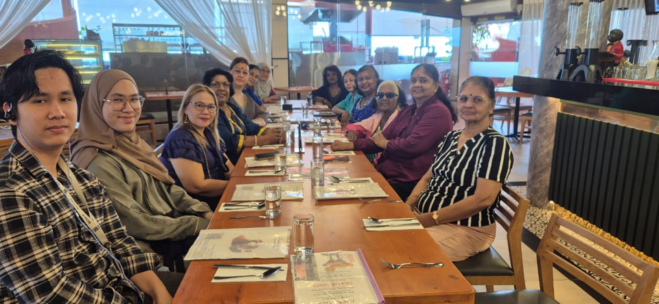 A group of people sitting around a long wooden table in a restaurant setting, smiling and facing the camera with menus, glasses, and cutlery arranged on the table.
Description generated by AI