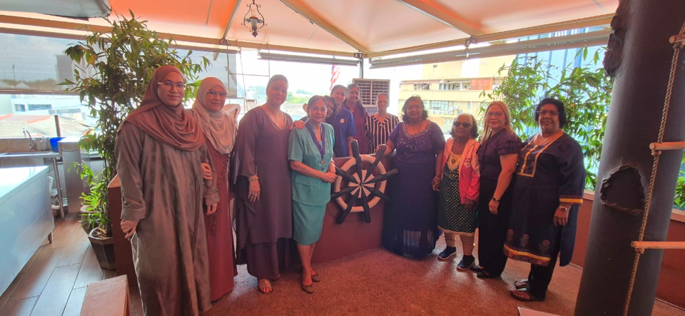 A group of people dressed in a mix of traditional and modern clothing stand together indoors near plants and a large decorative ship wheel.
Description generated by AI
