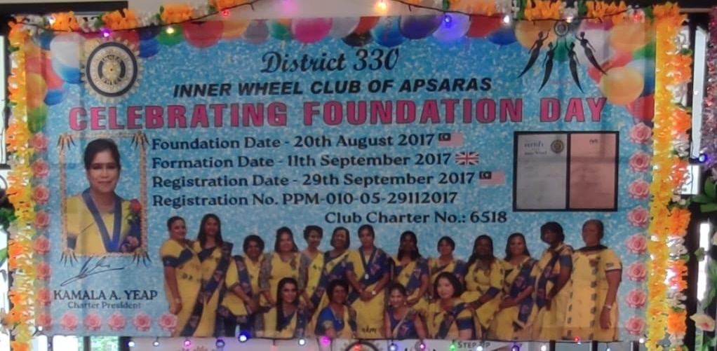 A decorated room with a banner celebrating the foundation day of the Inner Wheel Club of Apsaras featuring a group photo of people in matching yellow attire, a chair adorned with flowers and a sign reading "Charter President," and a table with a candle, documents, and string lights.
Description generated by AI