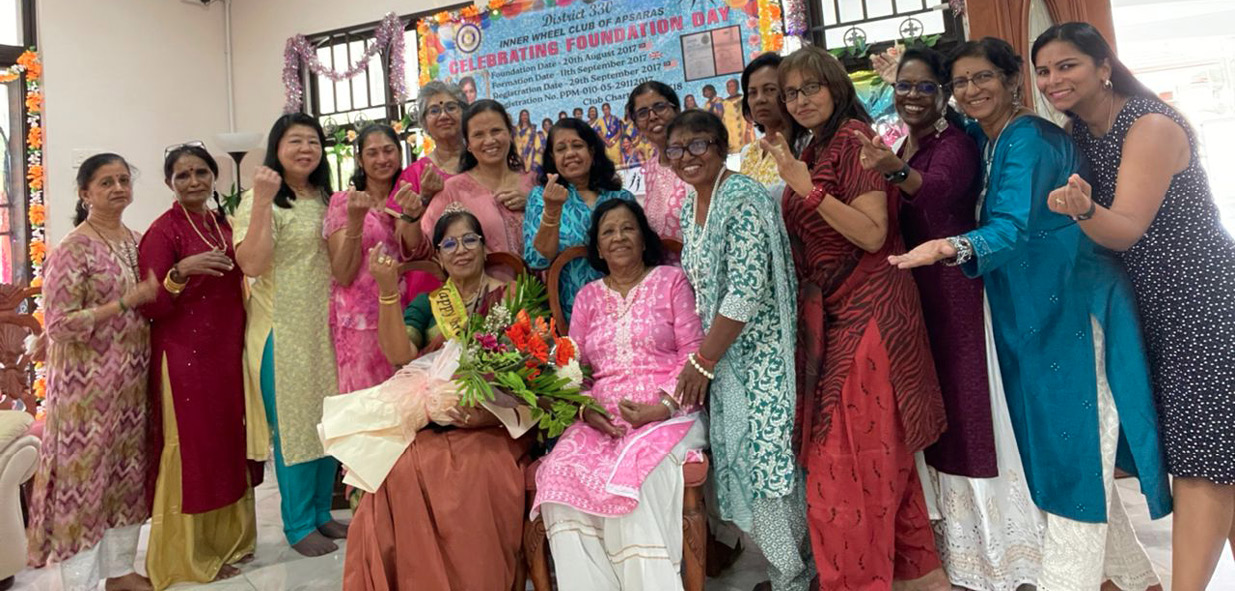 A group of people dressed in colorful traditional clothing are gathered indoors around two seated individuals holding a bouquet of flowers, celebrating an event with a decorated banner in the background.
Description generated by AI