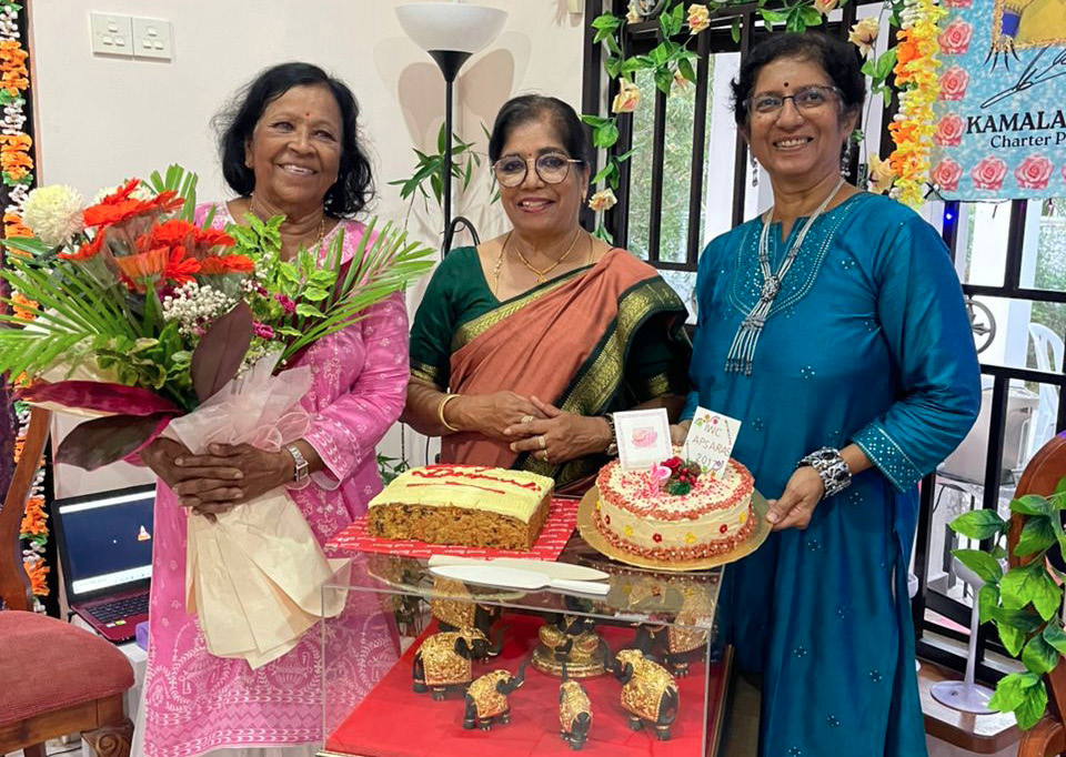 Three adults dressed in traditional clothing stand indoors smiling with one holding a bouquet of flowers and two decorated cakes placed on a table in front of them.
Description generated by AI