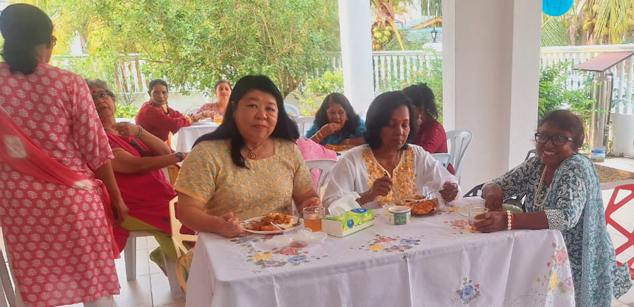 Several adults are seated around a table covered with a white embroidered tablecloth, engaging in an activity or conversation in a bright, open indoor space with red and black chairs.
Description generated by AI