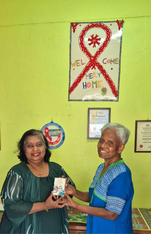 Two adults smiling and standing indoors near a desk, one wearing a green dress and the other in a blue patterned outfit, holding a small box together with a bright green wall and framed certificates in the background.
Description generated by AI