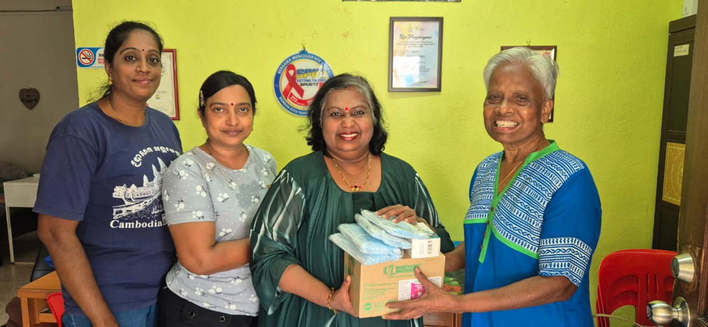 Four adults stand indoors against a green wall with framed items while one hands a box and packages to another, all smiling.
Description generated by AI