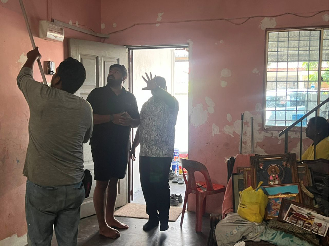 People are gathered inside a room with pink walls, one person is pulling a rope attached to the ceiling while others stand or sit nearby, and various household items and framed pictures are visible.

Description generated by AI