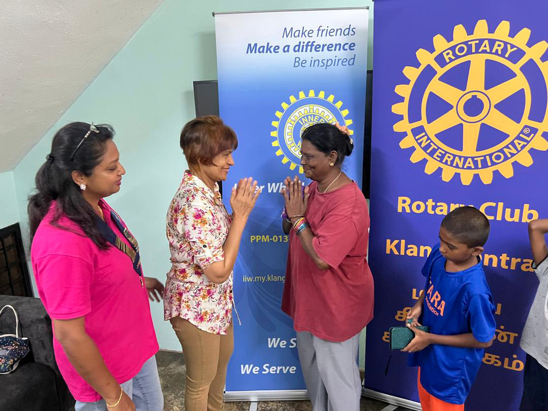 Four people stand in front of two Rotary Club banners, with two adults greeting each other with hands pressed together while another adult and a child look on.

Description generated by AI