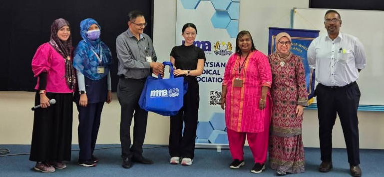 Seven adults stand in a row on a stage with a blue carpet, some wearing colorful traditional clothing, and one person holding a blue bag, with a whiteboard and banners in the background.
Description generated by AI