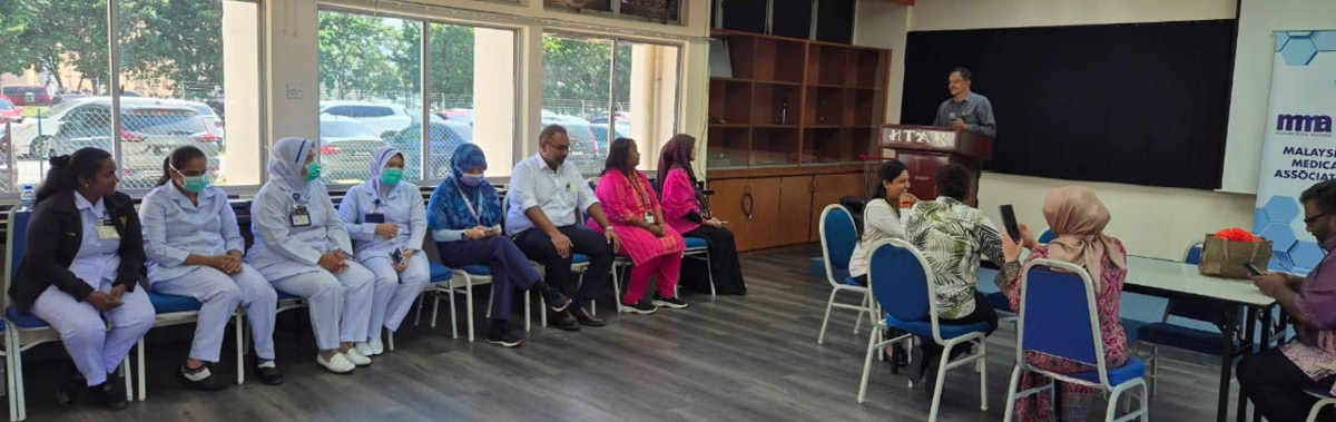 People seated in a room with large windows, some wearing medical scrubs and masks, listening to a person speaking at a podium.
Description generated by AI