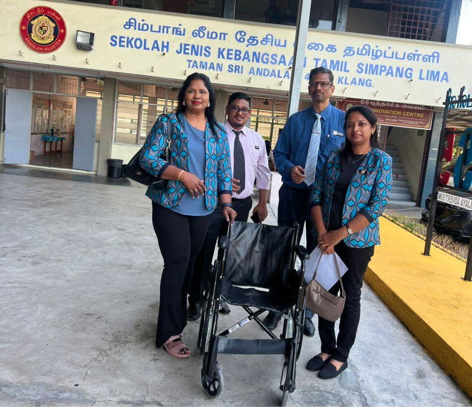Four adults standing around a wheelchair outside a building with a sign in multiple languages including Tamil and English.

Description generated by AI