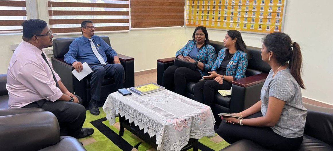 Five people dressed in business casual attire are seated in a modern office with large windows and a bulletin board, engaged in a conversation around a table covered with a lace cloth.

Description generated by AI