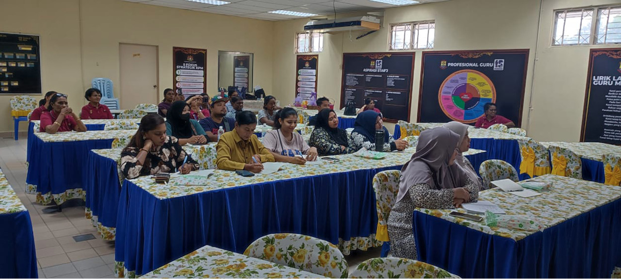 People seated at tables covered with floral and blue tablecloths in a classroom or meeting room setting with posters on the walls.
Description generated by AI