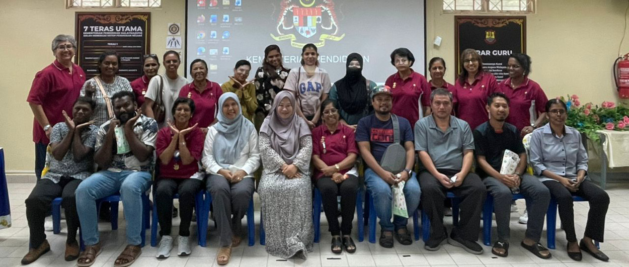 A group of people posing for a photo in a room with a projector screen displaying a crest and text, with some seated on blue chairs and others standing behind them.
Description generated by AI