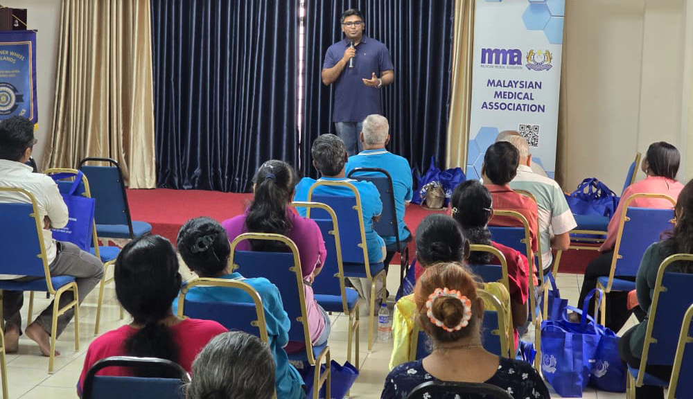A person wearing glasses and a dark blue shirt is speaking into a microphone on a small stage in front of an audience seated in rows of chairs in a conference room with a banner for the Malaysian Medical Association.
Description generated by AI