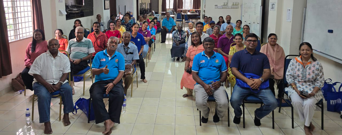 A group of adults seated in rows inside a well-lit room with tiled floors, some wearing blue shirts and others in casual clothing, facing forward as if attending a meeting or class.
Description generated by AI
