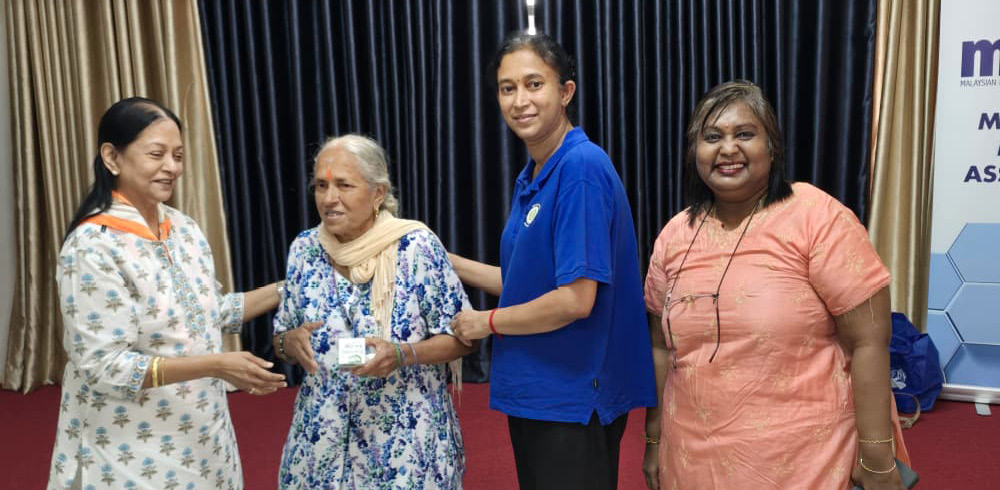 Four adults stand on a stage with dark curtains in the background, with one person in a floral dress receiving an item from another person in a white and orange patterned outfit, while two others in blue and peach clothing stand nearby smiling.
Description generated by AI