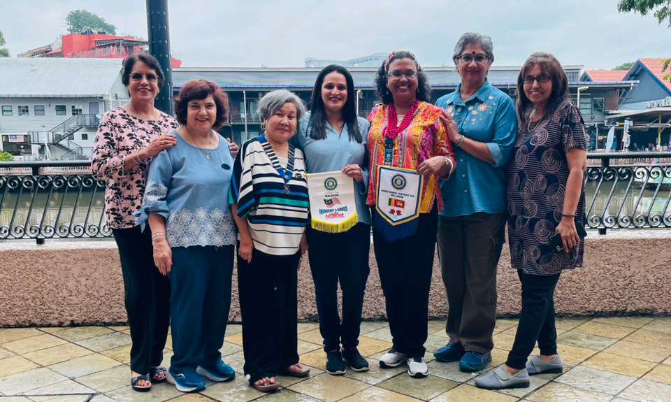 Six adults standing side by side on a tiled patio near a waterfront, with two holding colorful banners and smiling under a cloudy sky.
Description generated by AI