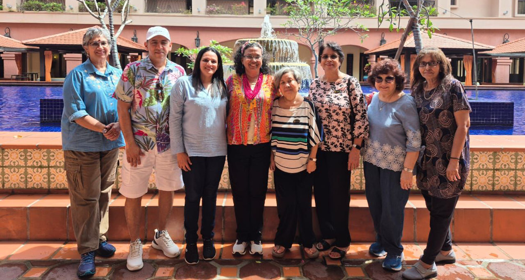 A group of eight adults standing side by side in front of a fountain and a building with balconies, with some wearing casual and colorful clothing and one person wearing a white cap.
Description generated by AI