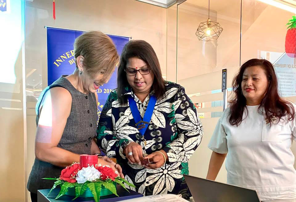 Three adults stand around a table with a laptop and a projector, with two of them focused on a decorative item with flowers and a candle.

Description generated by AI
