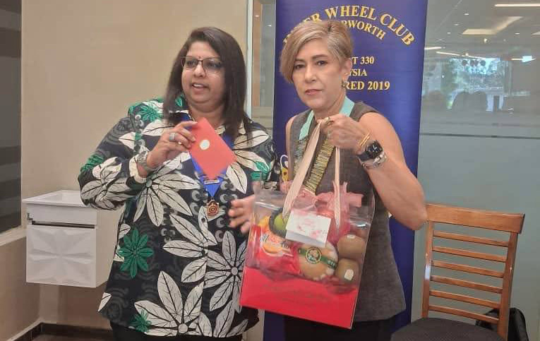 Two adults stand indoors near a banner, one holding a red gift bag filled with various items and the other holding a red envelope, with a festive decoration hanging above them.
Description generated by AI