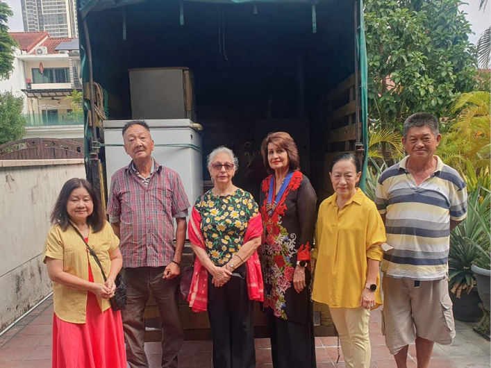Six adults stand together on a tiled outdoor area in front of a truck, dressed in casual and colorful clothing, with greenery and buildings in the background.
Description generated by AI