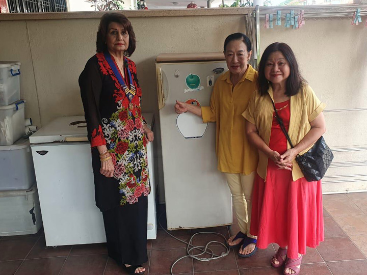 Three adults stand indoors near a refrigerator and freezer, with one person pointing at the refrigerator while the others stand close by, all dressed in colorful clothing.
Description generated by AI