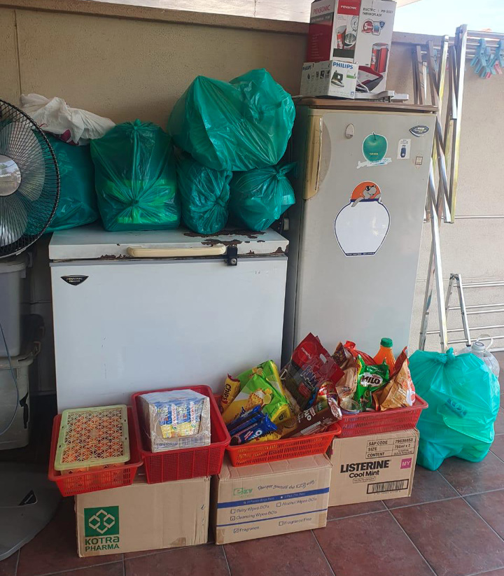 A cluttered corner with a white freezer, a white refrigerator decorated with stickers, several green plastic bags on top and beside the appliances, and multiple baskets and boxes filled with various packaged snacks and items on the floor.
Description generated by AI