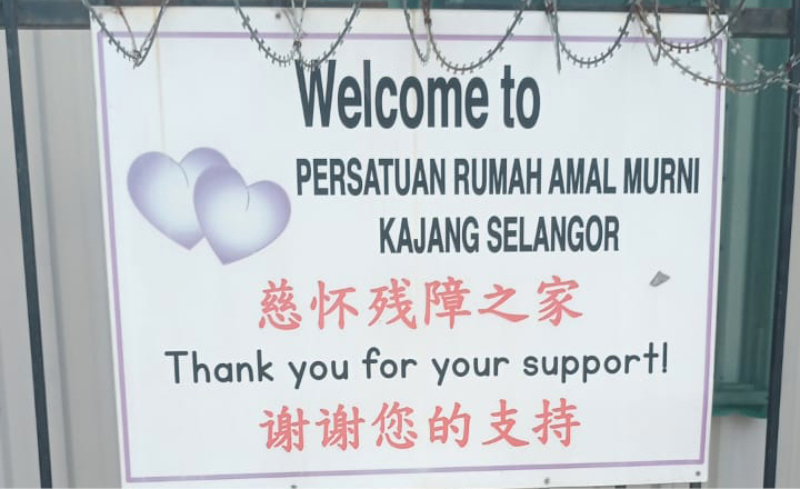 A sign on a metal fence welcomes visitors to Persatuan Rumah Amal Murni Kajang Selangor and thanks them for their support in English and Chinese, with two heart graphics and plants in pots below.
Description generated by AI