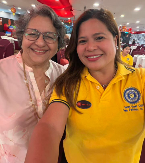 Two people smiling and posing for a selfie in a brightly lit indoor setting decorated with red lanterns, with one person wearing glasses and a light-colored top and the other wearing a yellow polo shirt with a Rotary Club logo.
Description generated by AI