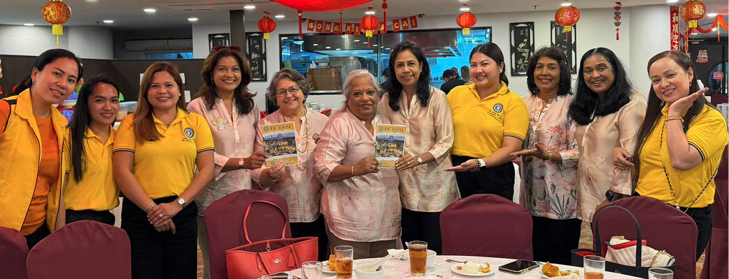 A group of people wearing yellow and light pink tops are standing together smiling behind a round dining table with plates of food and glasses of tea in a festive decorated room.
Description generated by AI