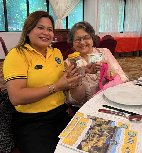 Two people sitting at a table in a room with large windows and curtains are smiling and holding up packaged products while one person wears a yellow polo shirt and the other wears glasses and light-colored clothing.
Description generated by AI