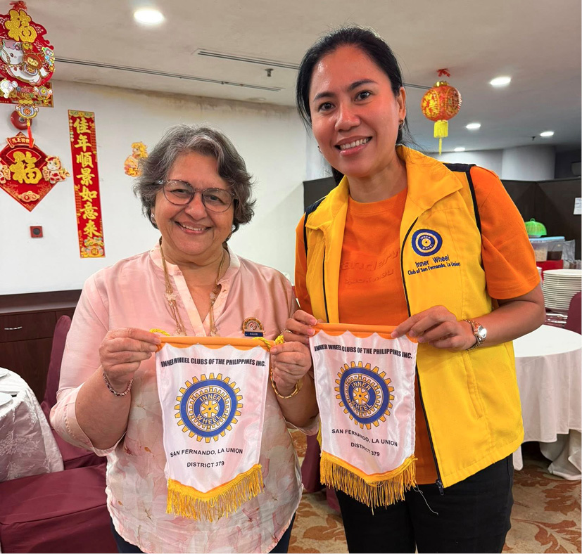 Two adults smiling and holding small banners with Rotary Club logos in a decorated indoor setting.
Description generated by AI