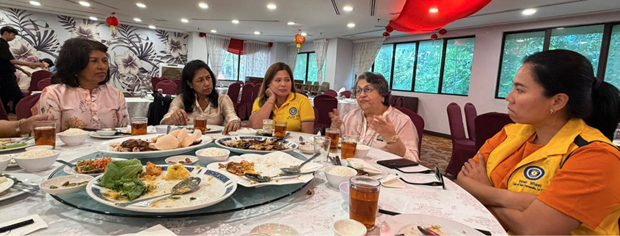 Five adults sit around a round table with plates of food and drinks in a decorated room, engaged in conversation.
Description generated by AI
