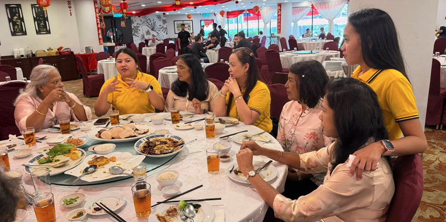 A group of people wearing casual clothing are seated around a round table in a decorated restaurant, engaging in conversation and sharing a meal with various dishes and drinks on the table.
Description generated by AI
