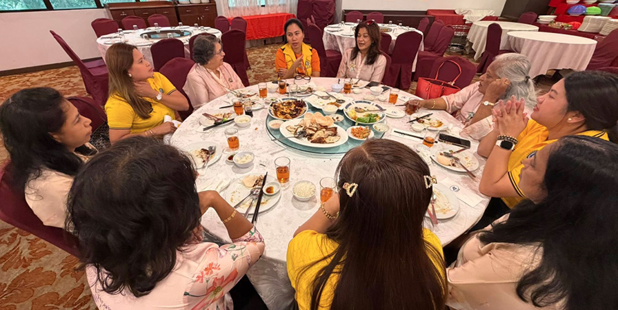 A group of people wearing yellow and light-colored clothing are seated around a round table with plates of food and drinks in a decorated dining room.
Description generated by AI