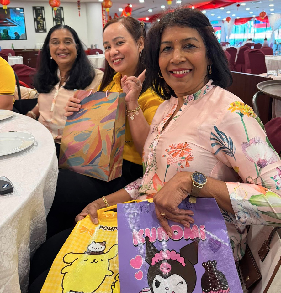 Three adults sitting at a round table in a decorated indoor setting, smiling and holding colorful gift bags.
Description generated by AI