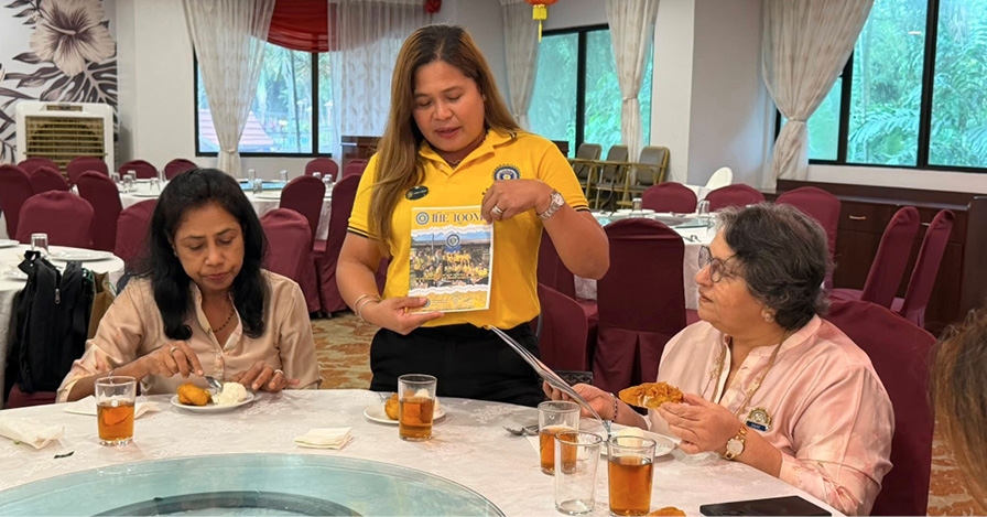 Three adults are seated around a round table with food and drinks in a restaurant, with one standing and holding a booklet while the others listen.
Description generated by AI