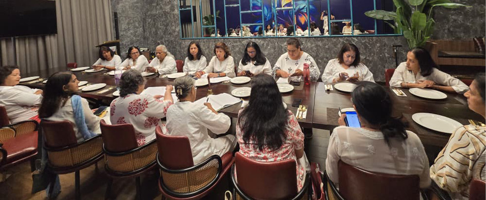 A group of people dressed in white are seated around a large dining table in a modern room with circular pendant lights and a decorative wall mirror.
Description generated by AI