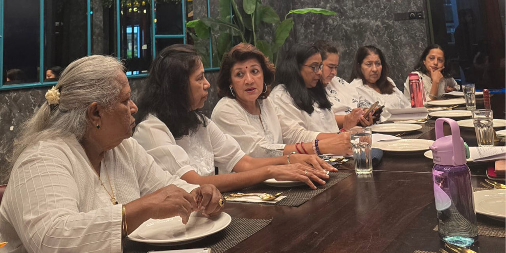 A group of adults dressed in white shirts are seated around a dark wooden table engaged in conversation with plates, glasses, and water bottles in front of them in a modern indoor setting.
Description generated by AI