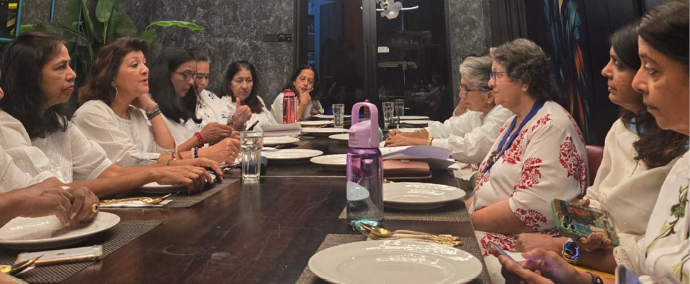 A group of adults seated around a long dining table in a modern room with dark walls, engaged in conversation and holding various items.
Description generated by AI