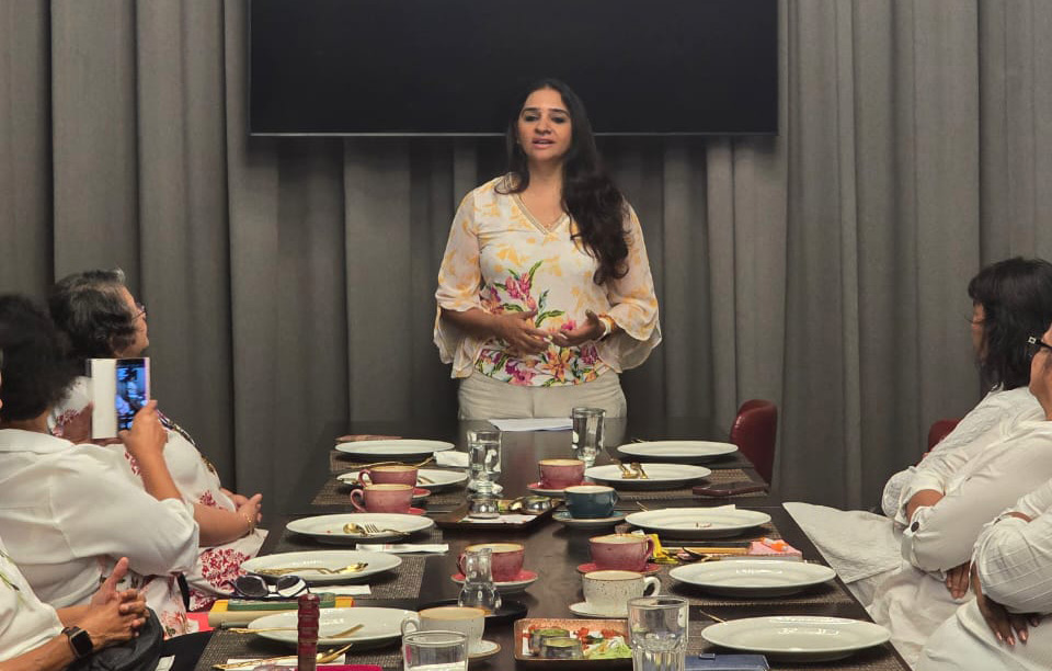 A person stands at the head of a dining table set with plates, cups, and glasses while several people sit on either side, listening attentively in a room with gray curtains and a modern circular light fixture above.
Description generated by AI
